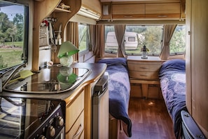A compact RV interior with a kitchenette, a bed with a patterned bedspread, and a window with curtains. - Caravan 'Vue Sur L'étang' with Lake View, Shared Pool and Private Terrace (Champsac)