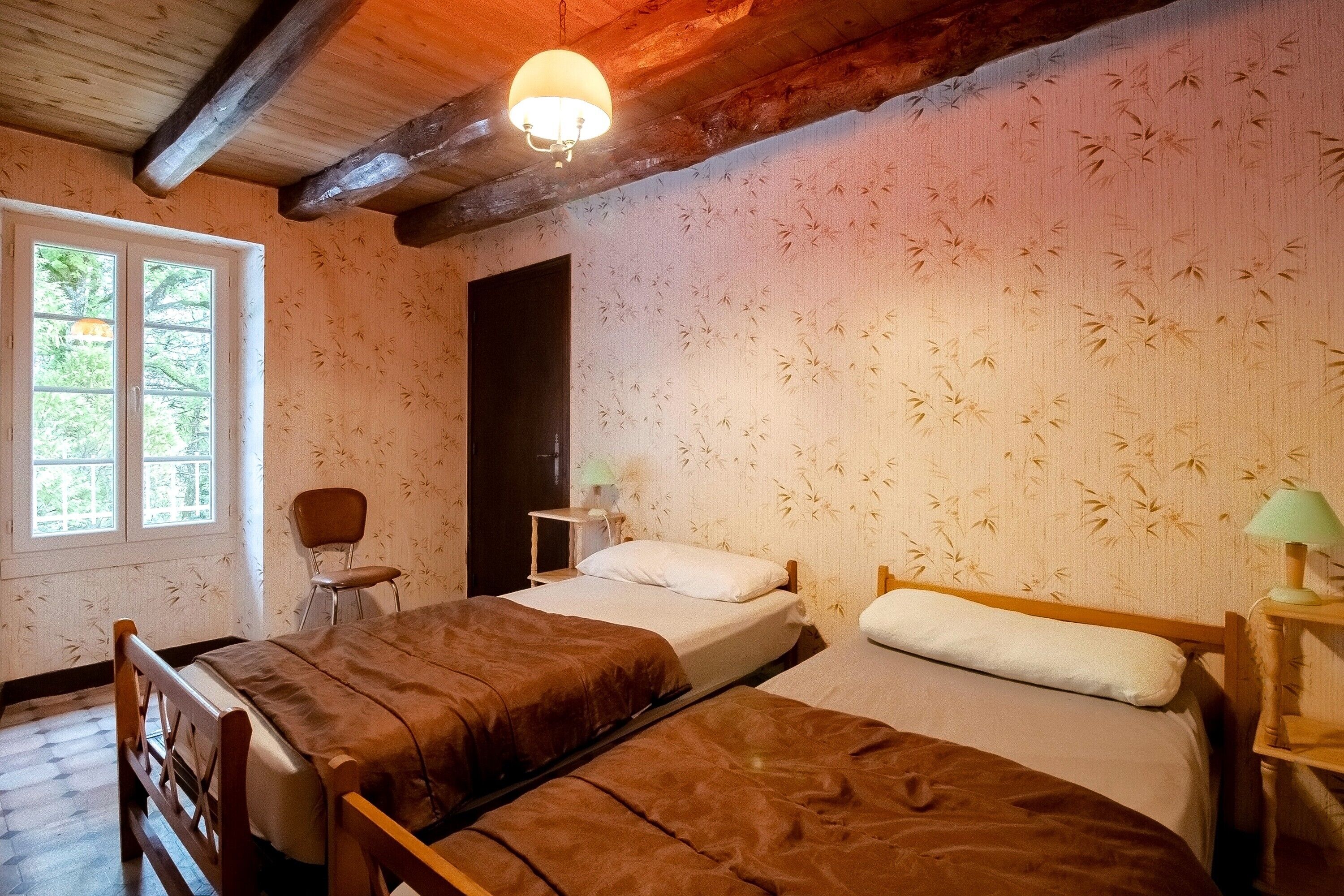 A room with two beds, a chair, a desk, a lamp, and a window with a view of greenery.