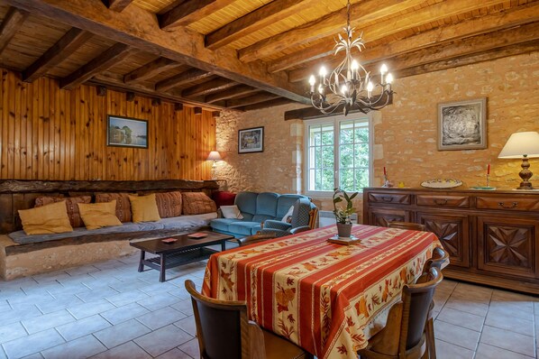 Un interior rústico con vigas de madera a la vista, una pared de piedra, un sofá, una mesa de comedor con mantel floral y una araña.