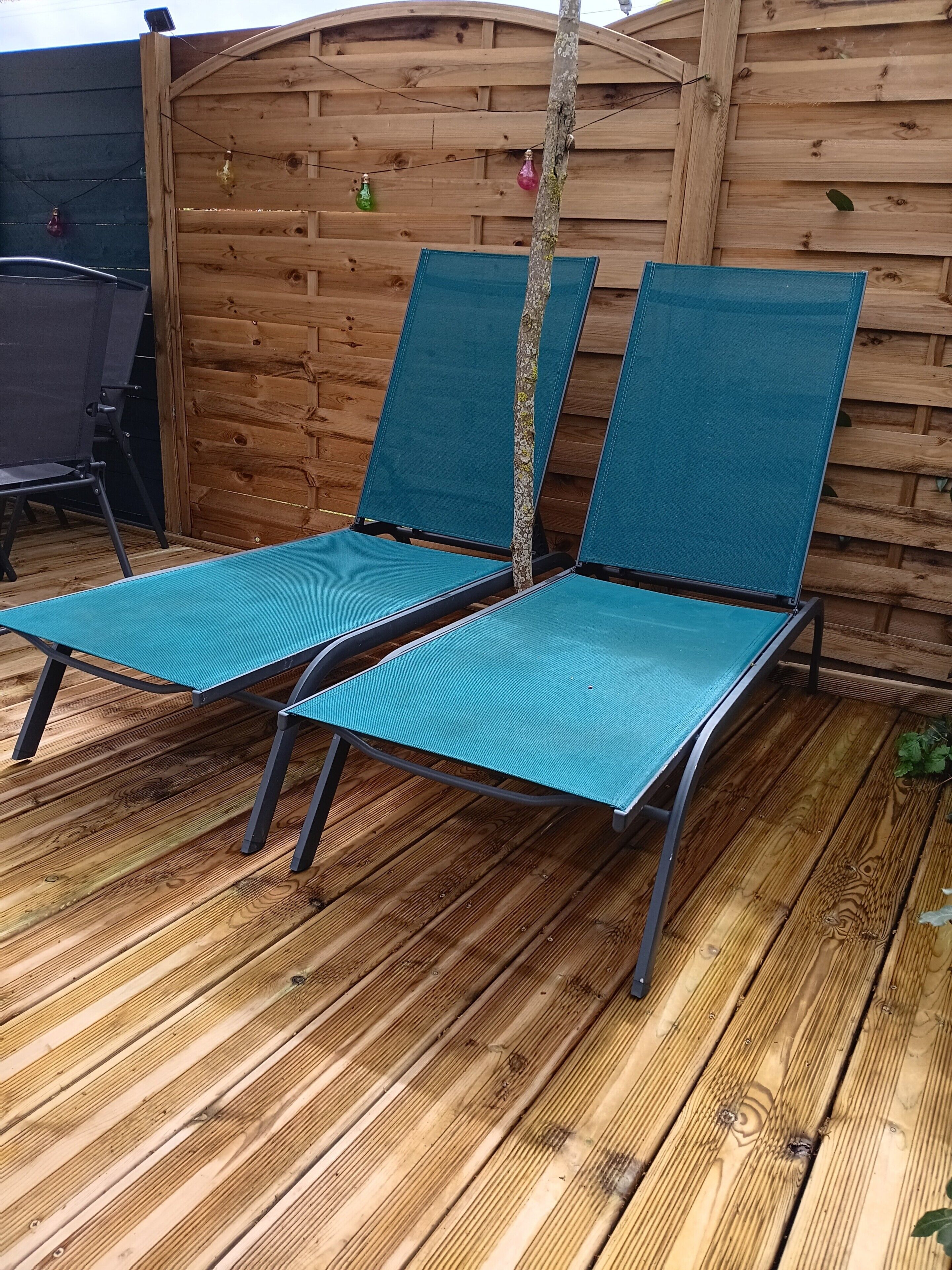 Två solstolar med metallramar och blått tyg, placerade på ett trädäck med ett trästaket i bakgrunden.