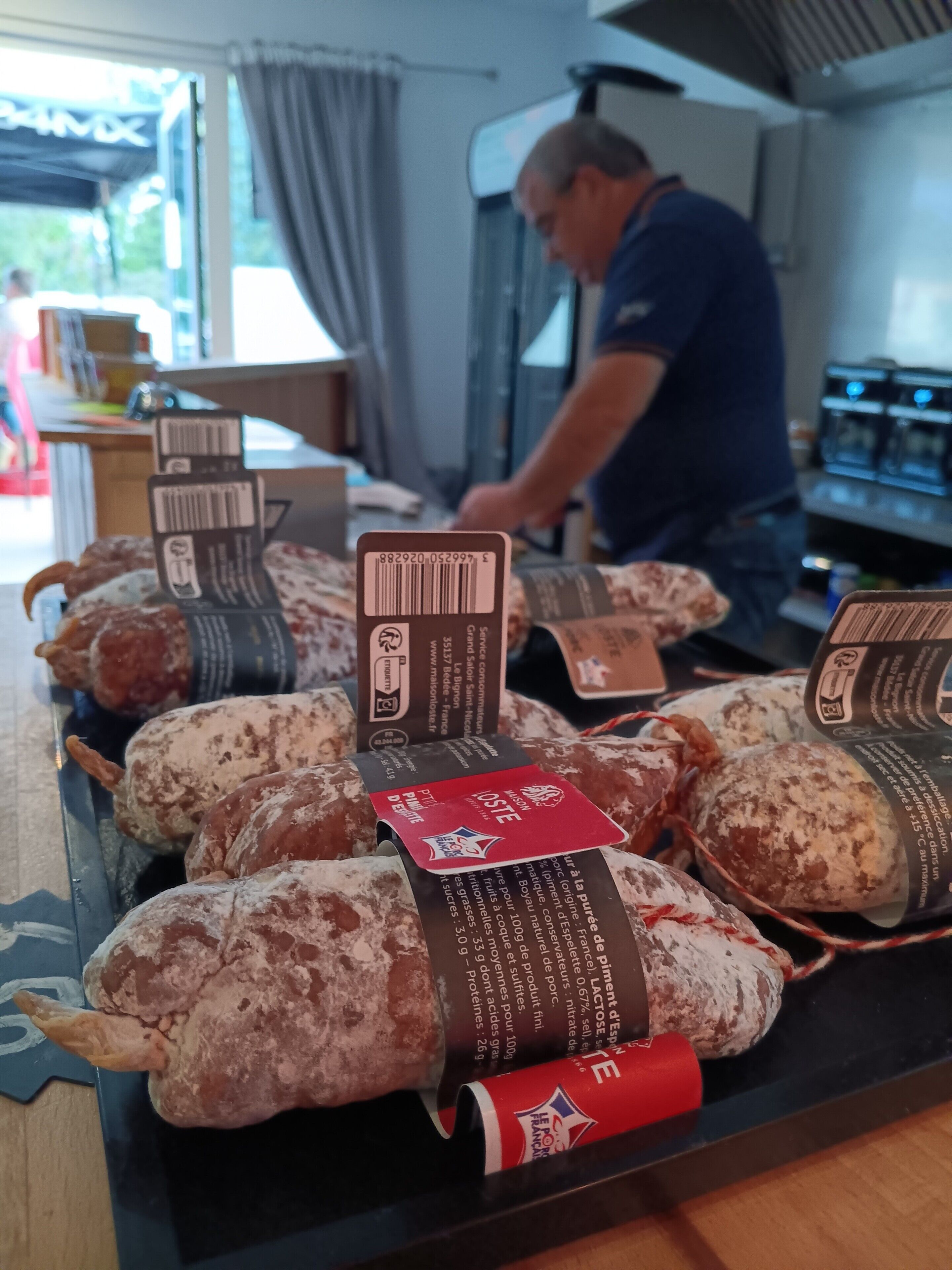 Een presentatie van gerookt vlees met etiketten, een persoon op de achtergrond en een raam met gordijnen.
