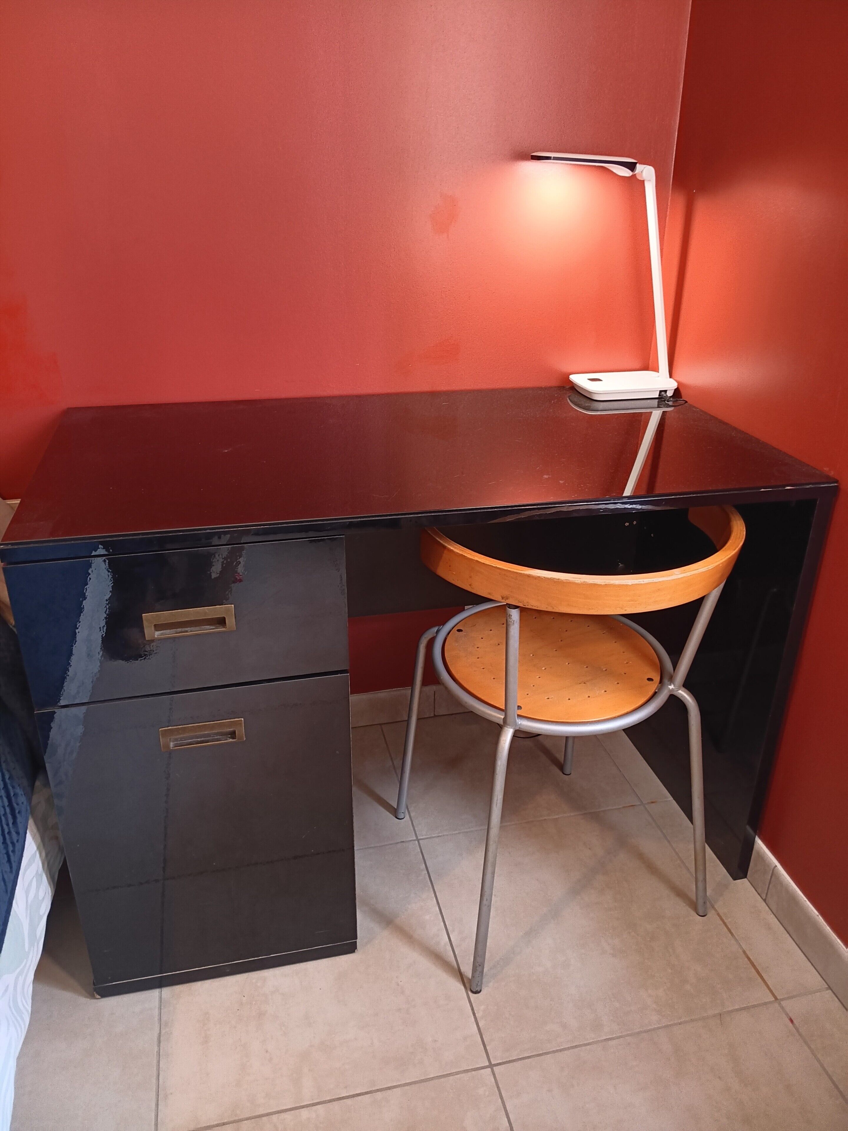 A black desk with a chair, a lamp, and a filing cabinet in a room with red walls and tiled flooring.