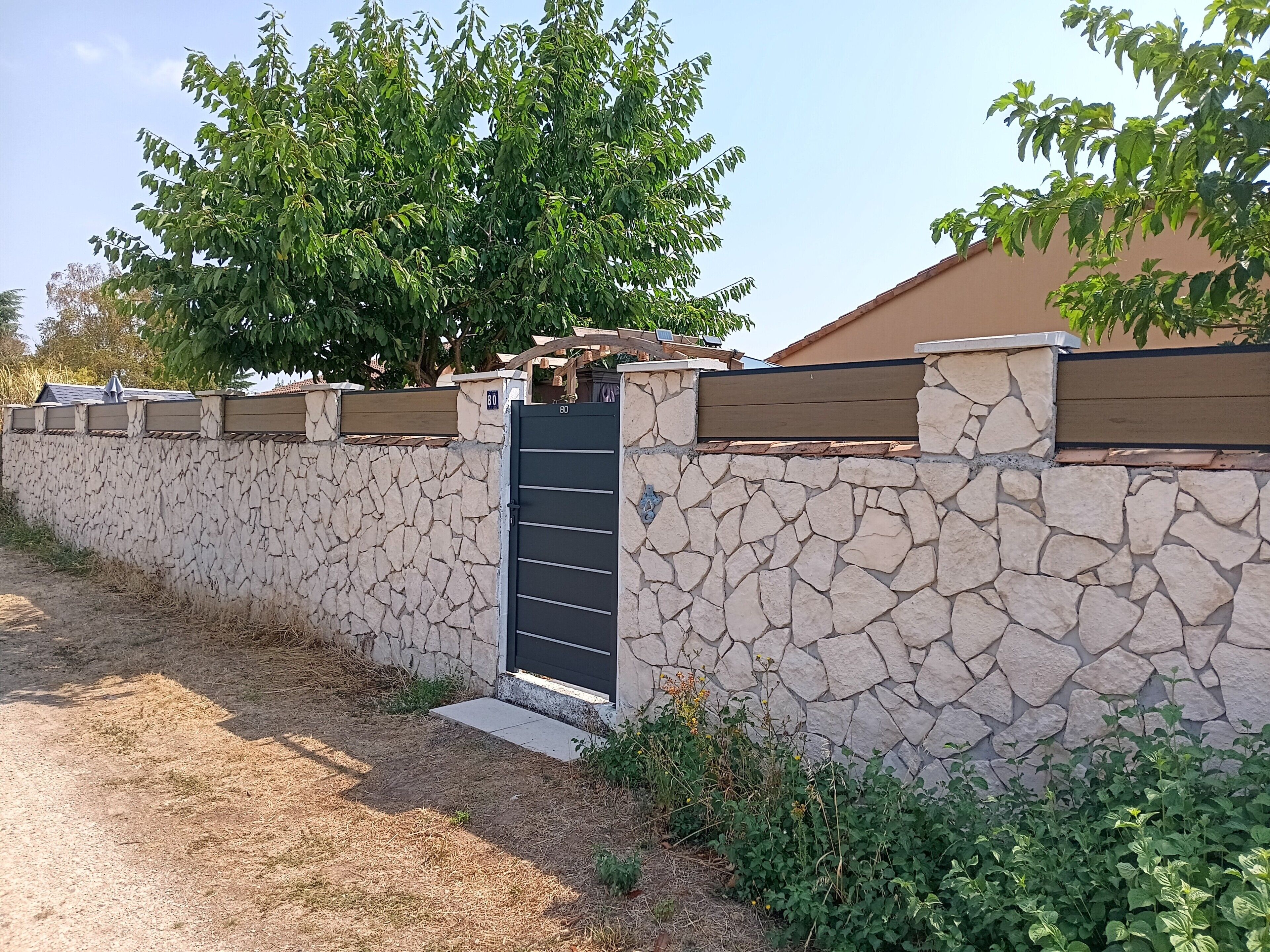 Un muro de piedra con una reja de metal, un árbol y un edificio al fondo.