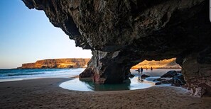 A beach with a large rock formation, calm water, and distant cliffs. - Holiday Home 'Casa Dayaday' with Mountain Views and Wi-Fi (Puerto del Rosario)