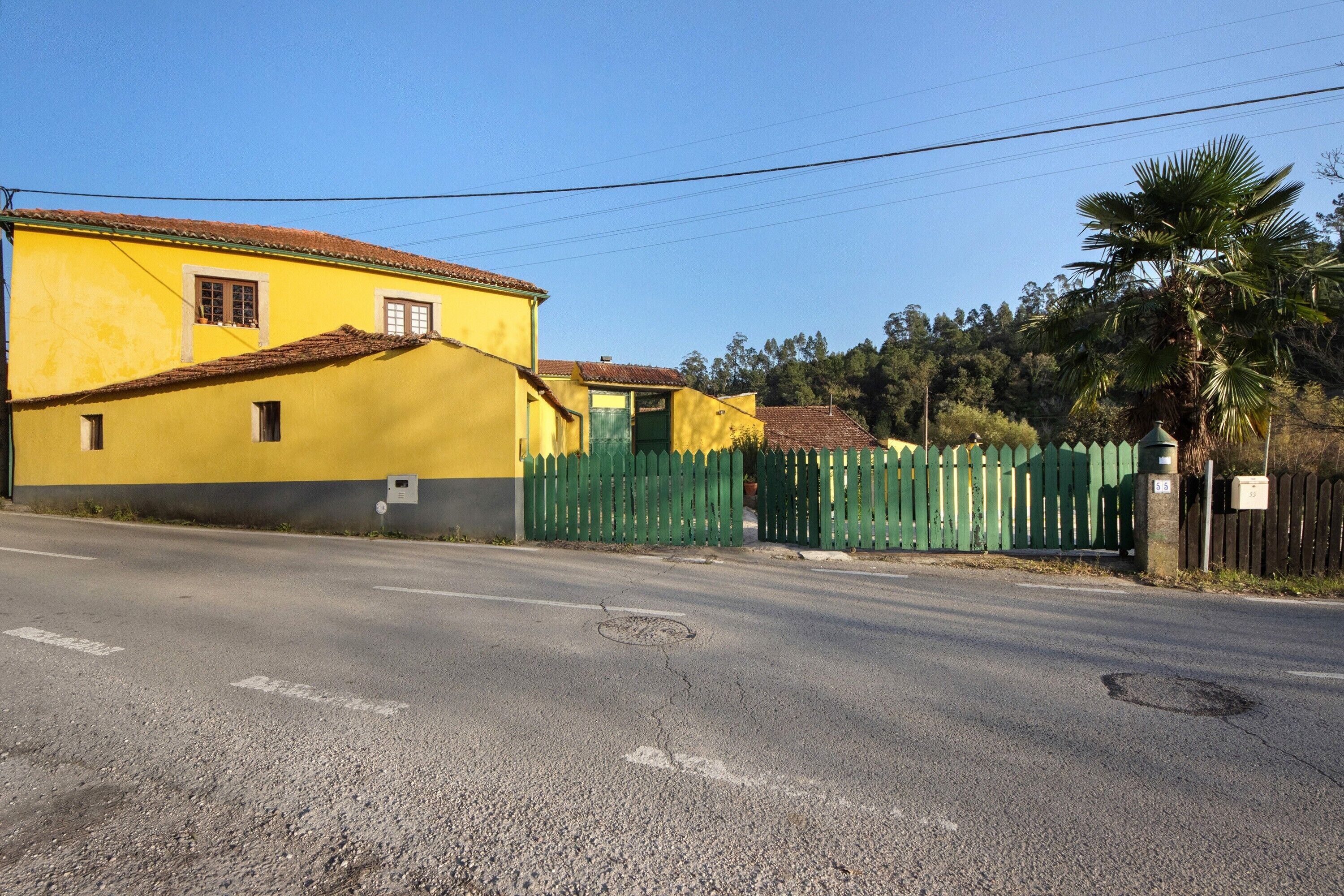 A yellow building with a green fence, a road, and a palm tree.