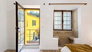 Een kamer met een schuifglasdeur die leidt naar een balkon, een klein raam met uitzicht op een geel gebouw en een bed met een bruine deken.