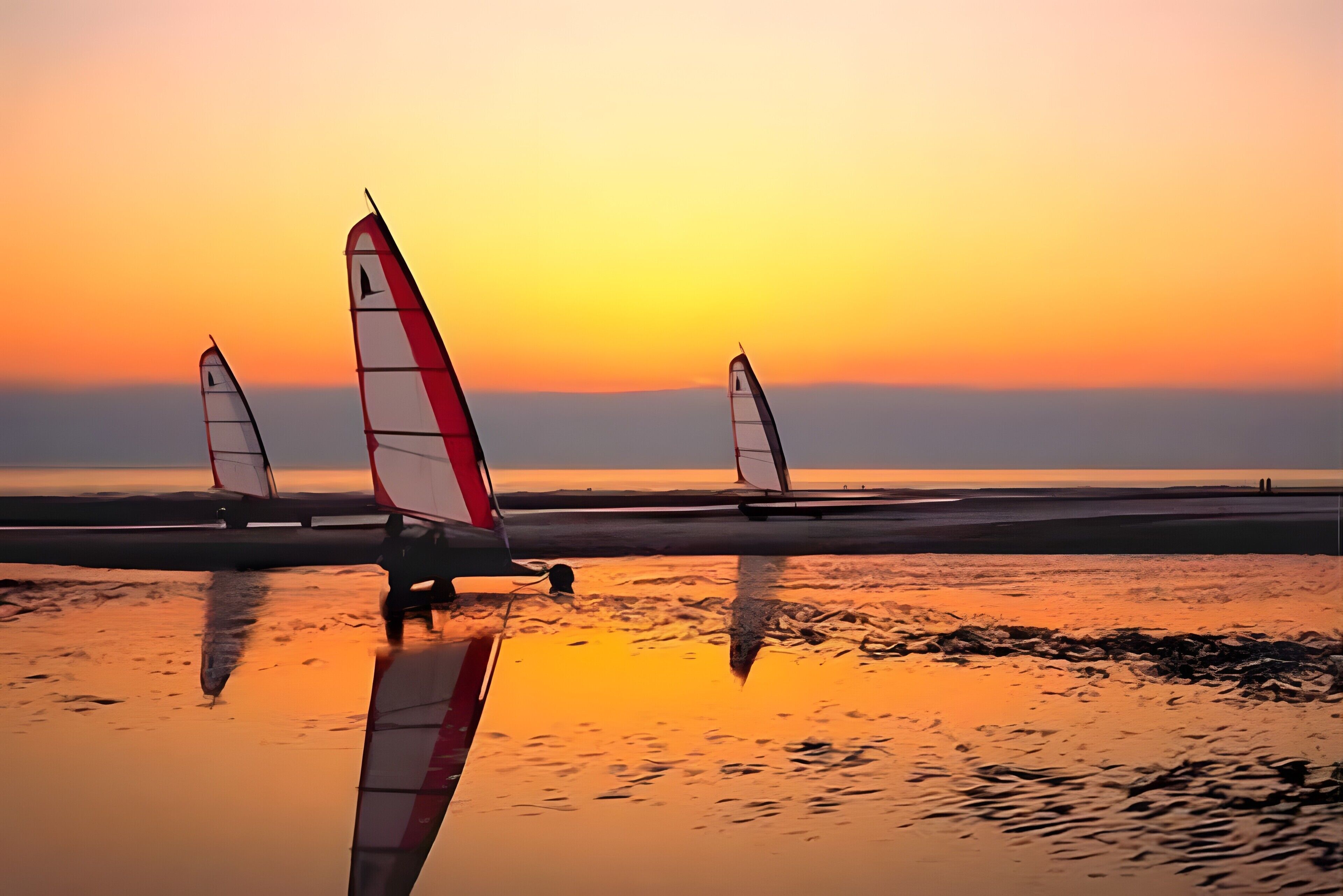 A beach with three sailboats and calm water reflecting the sunset.