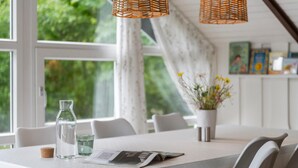 Un coin repas avec une table dressée pour deux personnes, une bouteille d'eau, un verre et un menu. Au-dessus de la table, on trouve des suspensions en osier, et sur le côté, un pot de fleurs avec des fleurs jaunes.