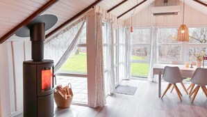 Une pièce chaleureuse avec un poêle à bois, de grandes fenêtres, un coin repas avec une table et des chaises, et un luminaire suspendu.
