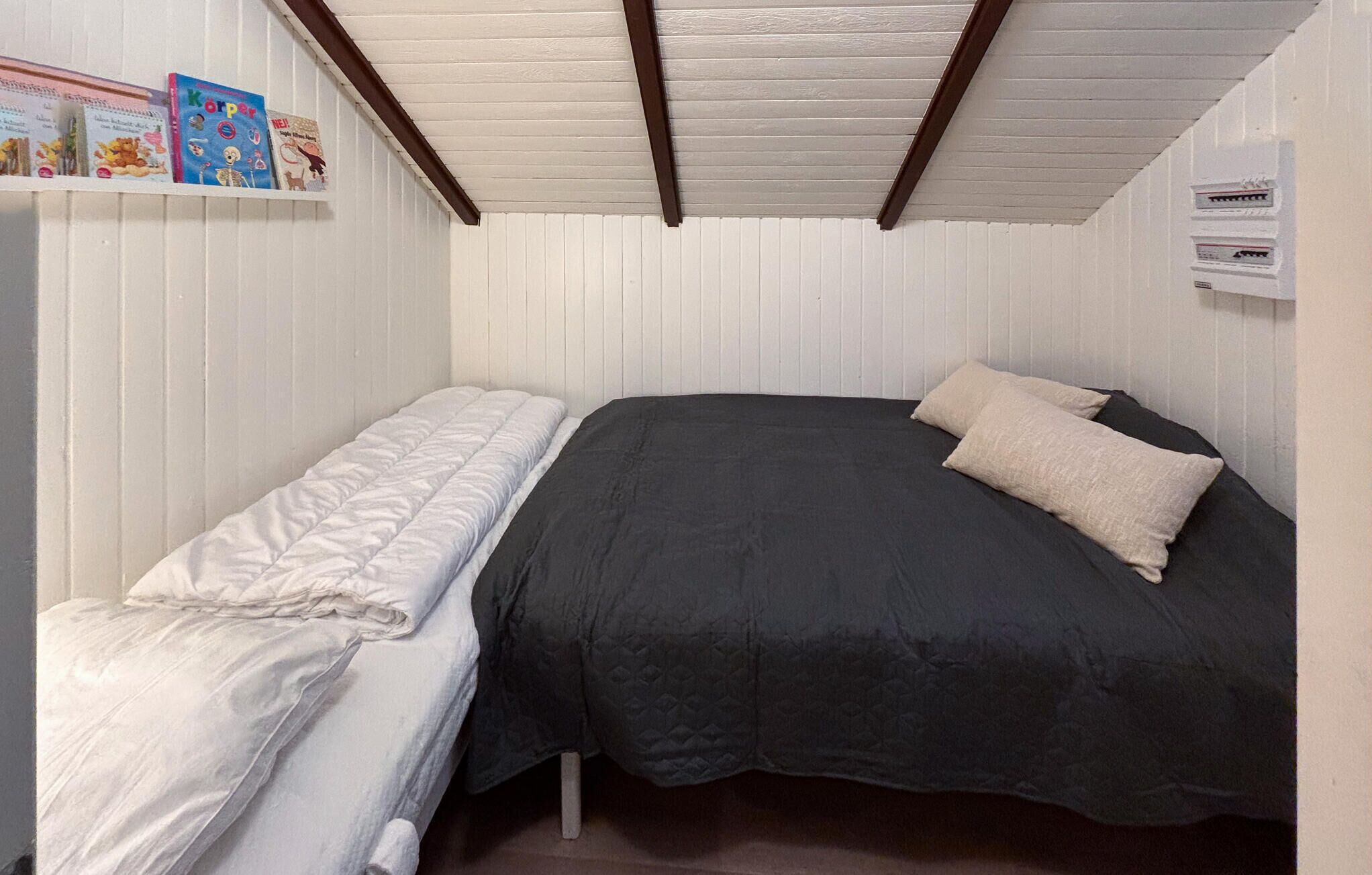 A small, single-bed room with a white headboard and a dark-colored bedspread. There is a shelf with books and a small poster on the wall.