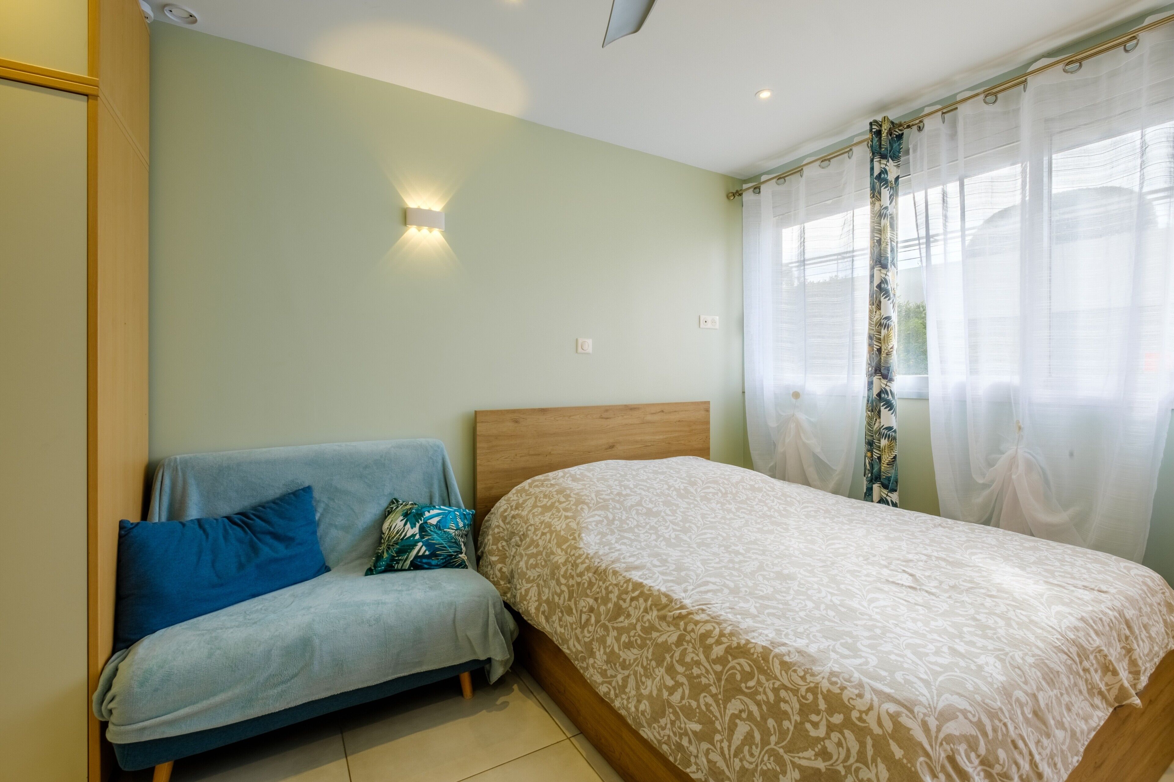 Une chambre à coucher avec un lit, un canapé, une lampe fixée au mur et des rideaux légers.