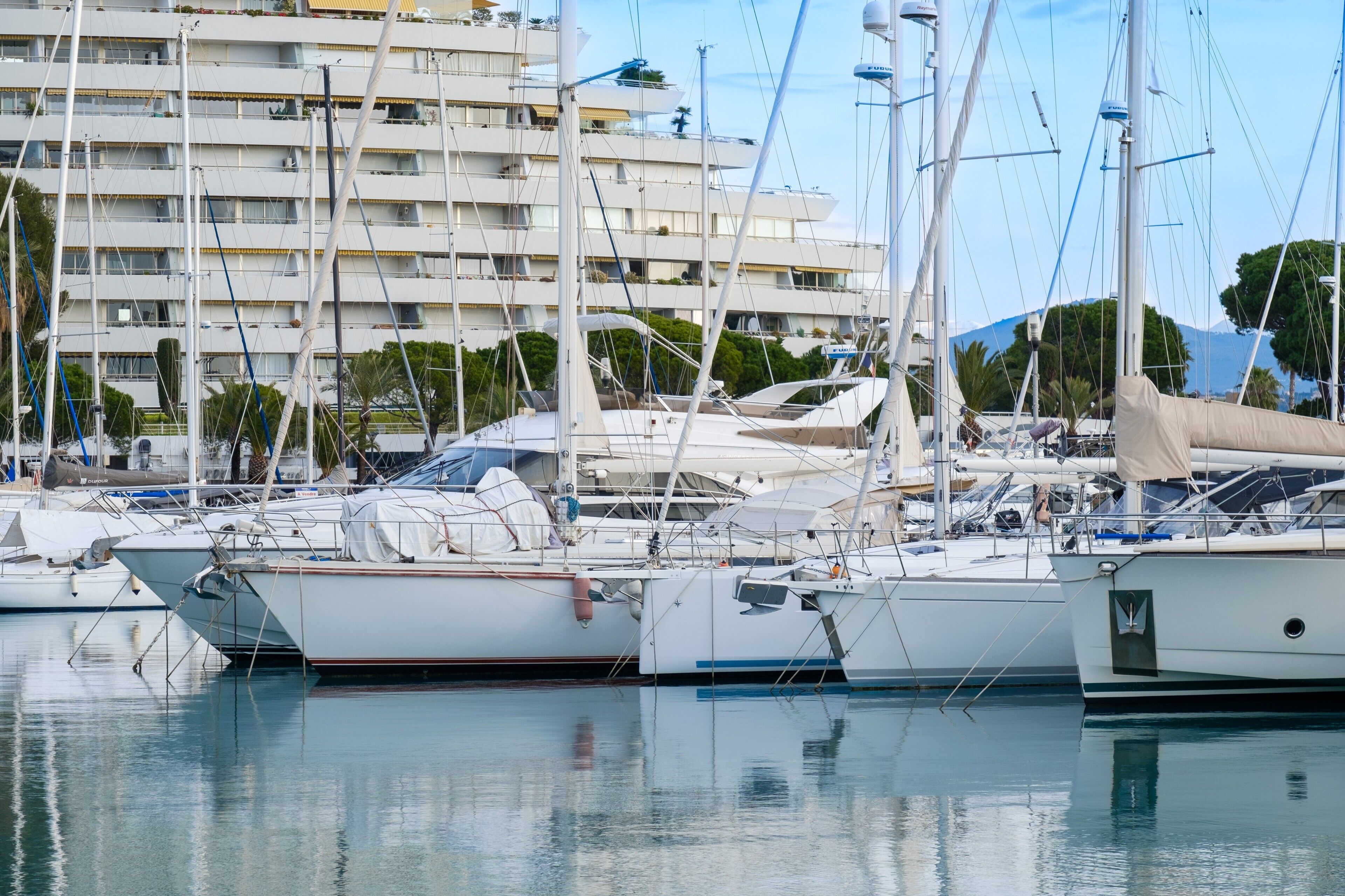 A marina with multiple boats docked, a building with balconies, and a clear sky.