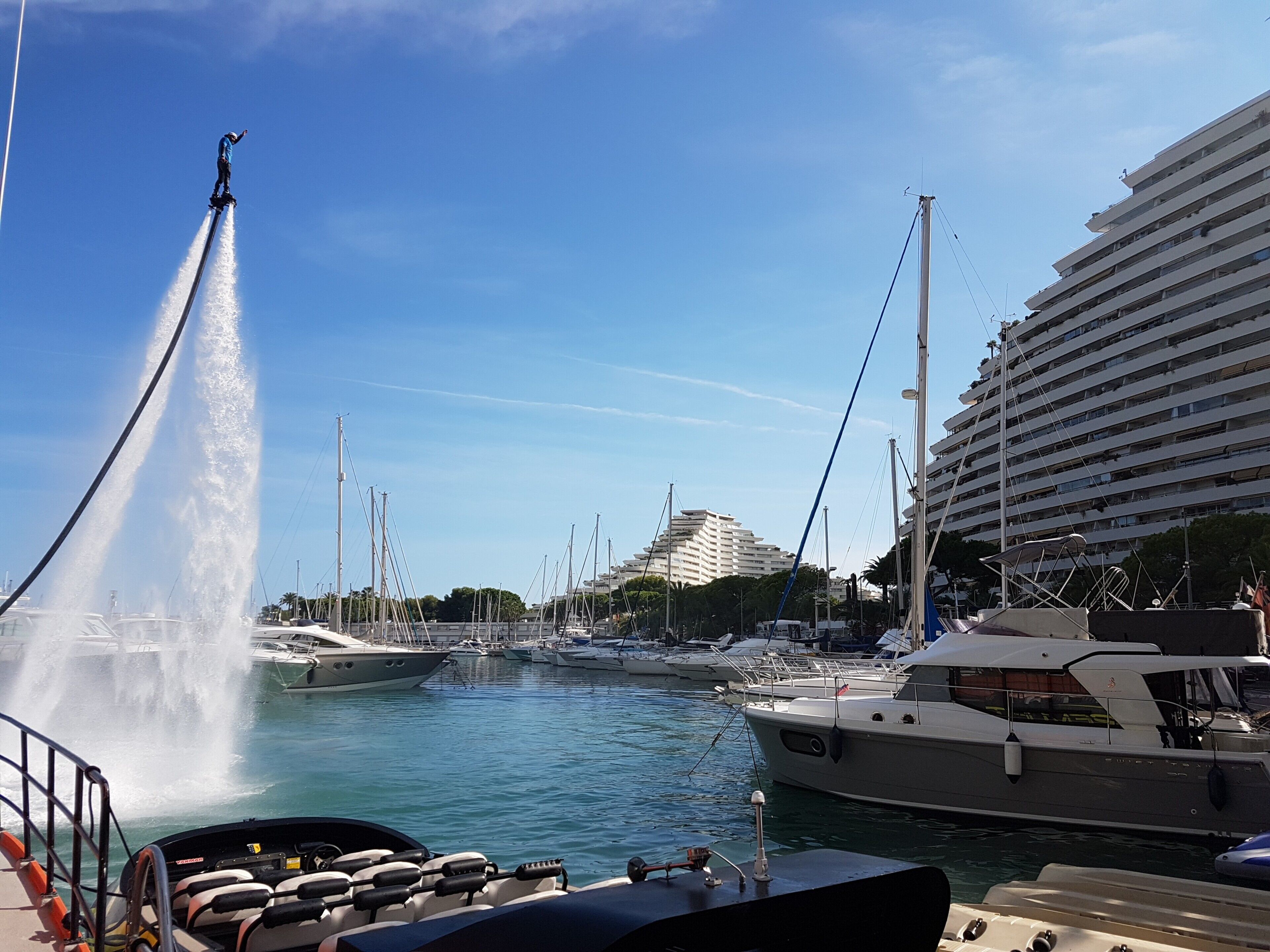 A marina with boats docked, a water fountain, and a large building in the background.