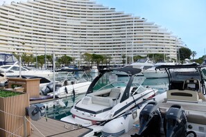 Un puerto deportivo con barcos amarrados, un gran edificio de hotel al fondo y una estructura de madera con el letrero 'NAUTIC EVASION'.