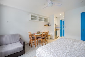 Una habitación con cama, sofá, mesa de comedor con sillas, ventilador de techo y una zona de cocina con puertas azules.