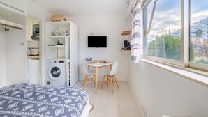 Une pièce de taille modeste comprenant un lit, un lave-linge, une petite table avec des chaises, un téléviseur et une fenêtre donnant sur un paysage agréable.