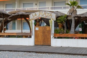 Un cancello in legno con un cartello che riporta la scritta 'LA PLAYA' sopra, che conduce a un edificio con un balcone e ombrelloni di paglia.