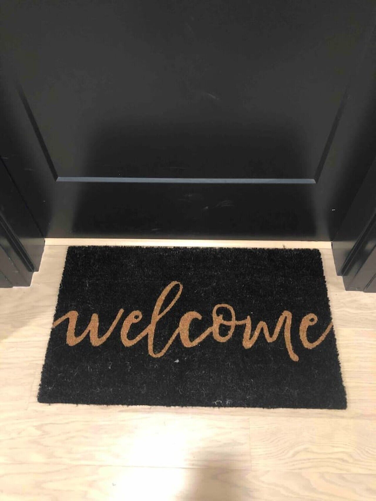 Un felp de porta negre amb la paraula 'welcome' escrita en lletres daurades amb cal·ligrafia.
