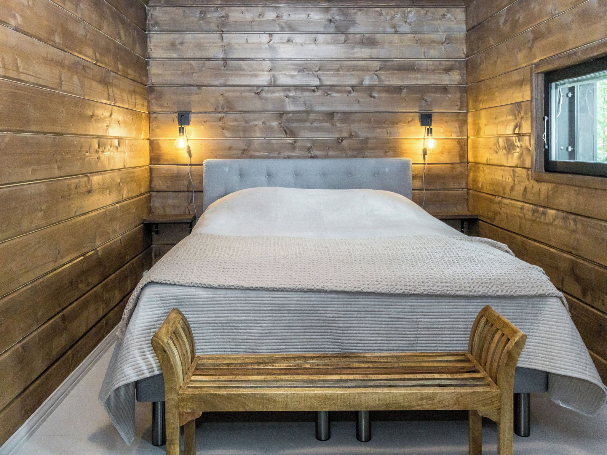 Una camera in una baita di legno con un letto, una panca e due luci fissate al muro.