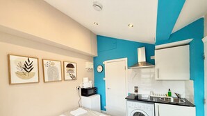 A compact hotel room with a bed, a washing machine, a kitchenette, and artwork on the wall.