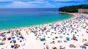 En fullsatt strand med många färgstarka parasoller och tält, ett klart blått hav och en byggnad på höger sida.