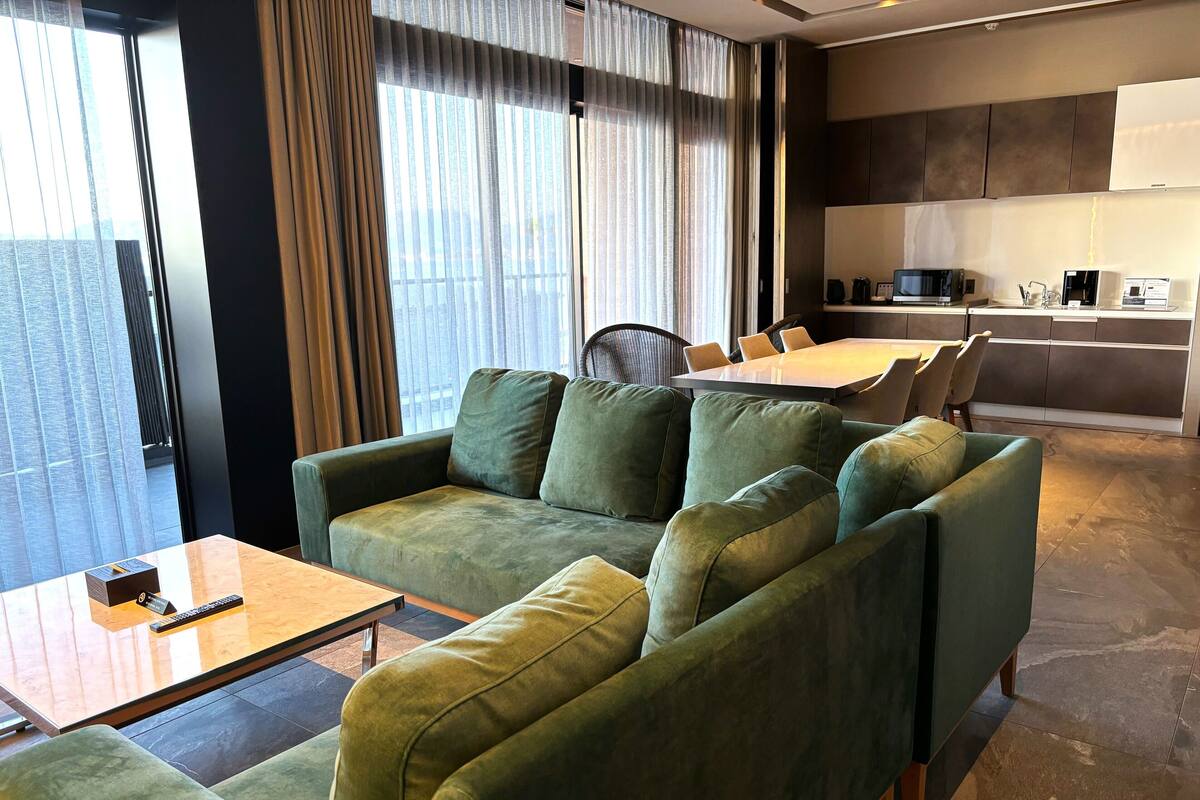 A modern living room with a green sofa set, a dining area with a table and chairs, and a kitchen area with appliances.