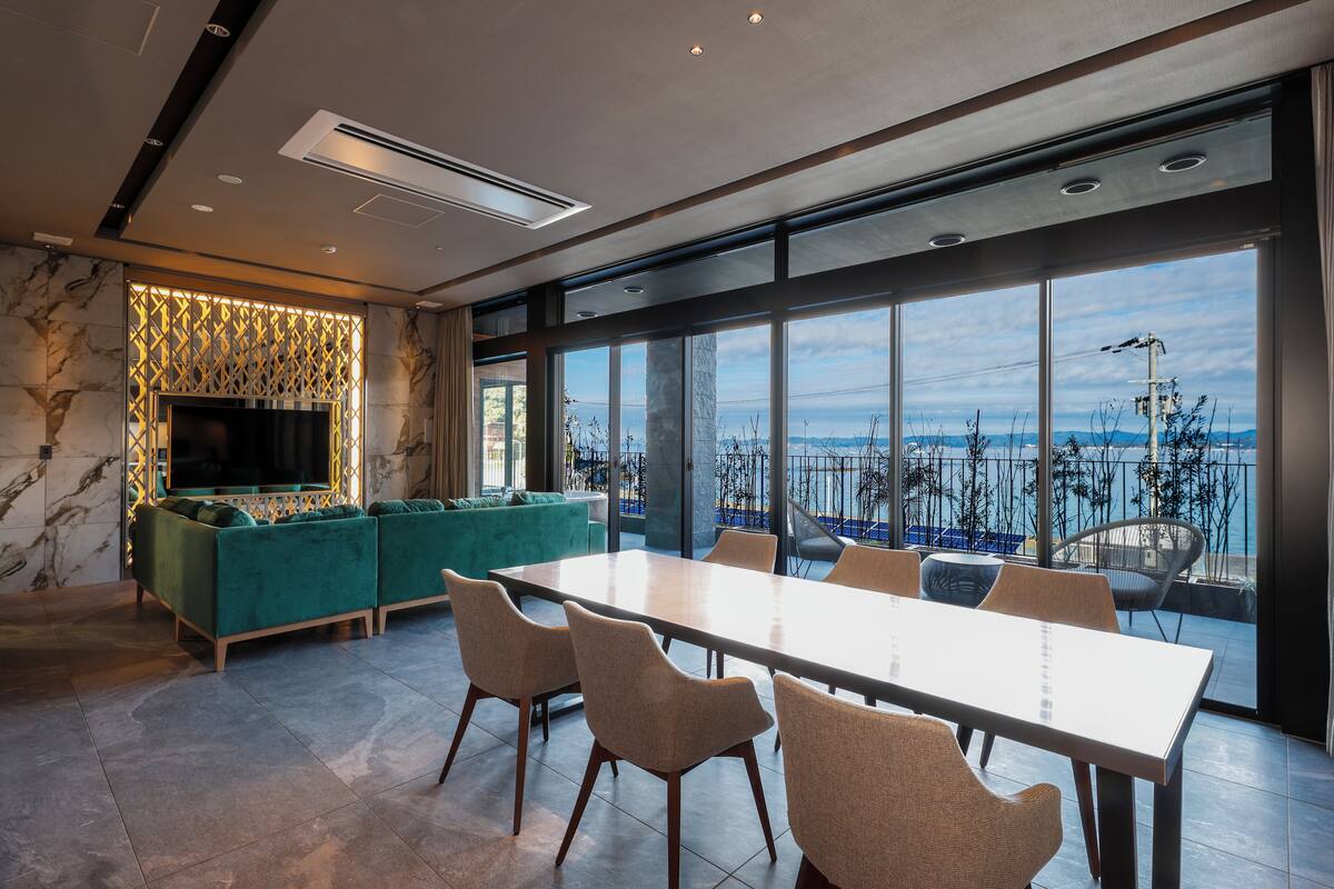 A modern interior with a large dining table, comfortable seating, a TV, and a view of the sea.