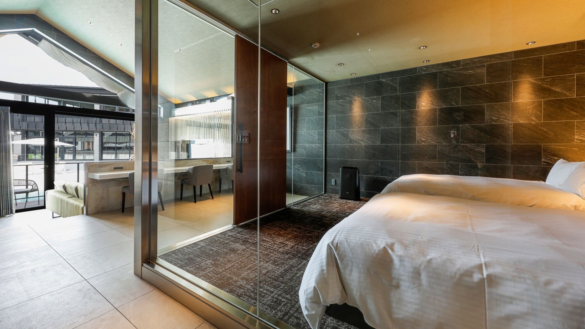 Ein modernes Hotelzimmer mit einem großen Bett, einer Glastrennwand, einem Essbereich und Blick ins Freie.