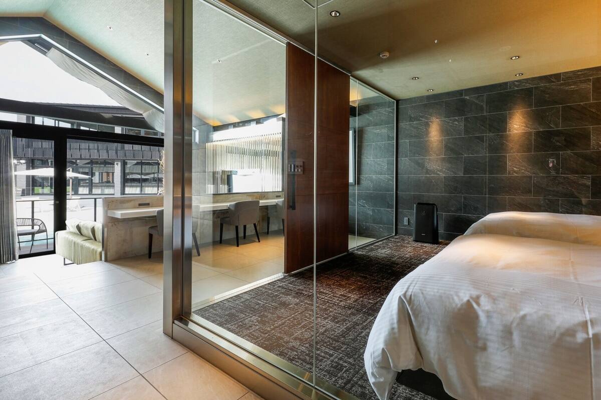 A modern hotel room with a large bed, a glass partition, a dining area, and a view of the outdoors.