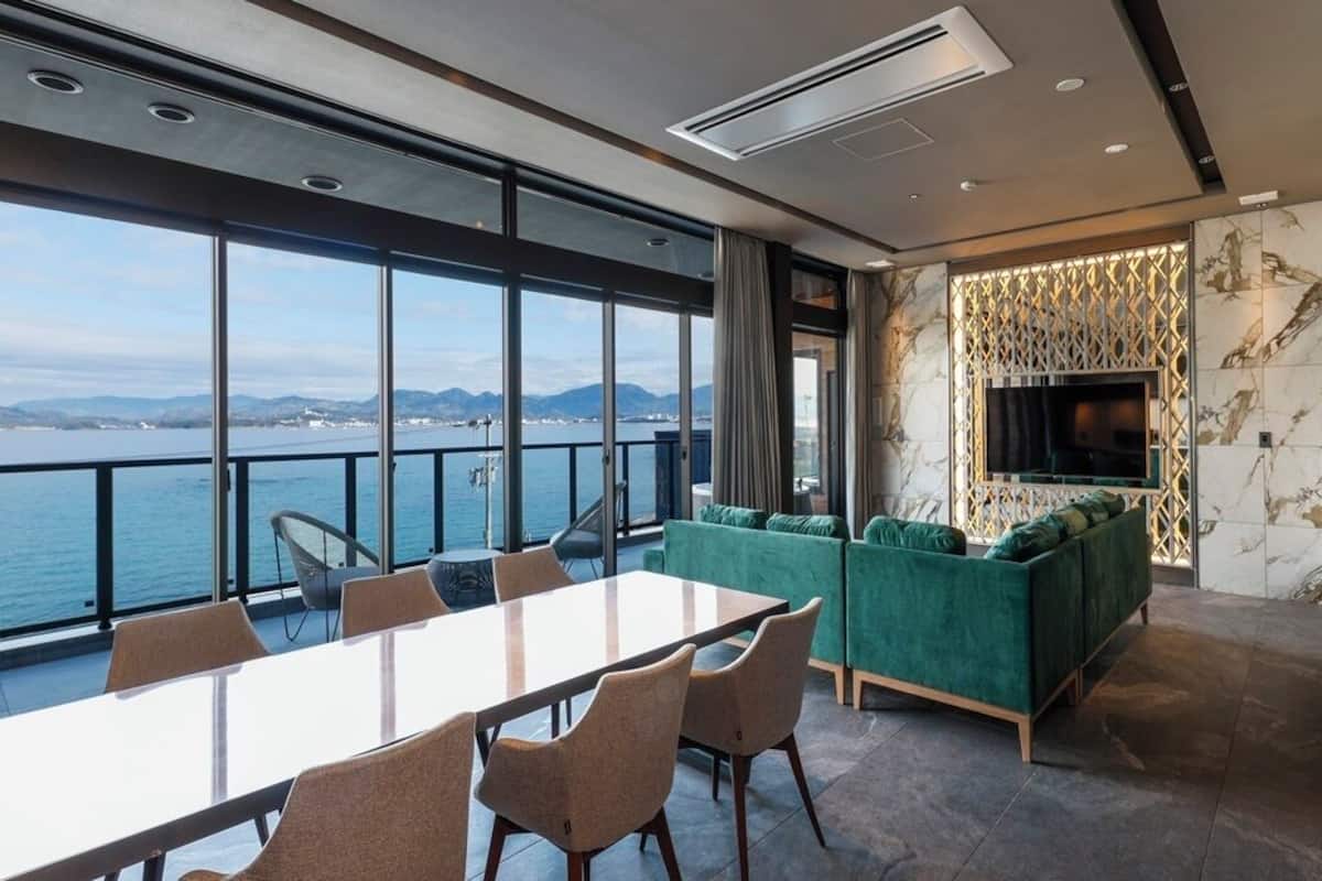 A modern room with a large table and chairs, a green sofa, a TV, and a view of the sea.