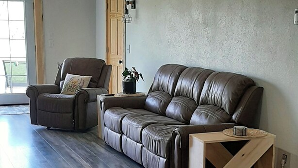 A spacious living room with a large leather sofa and a smaller armchair, wooden side table, and a wooden cabinet.