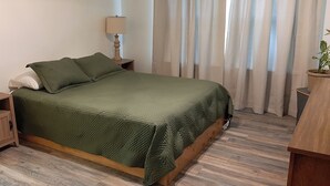 A neatly made bed with a green quilt, two bedside tables with lamps, a wooden nightstand, and a potted plant.