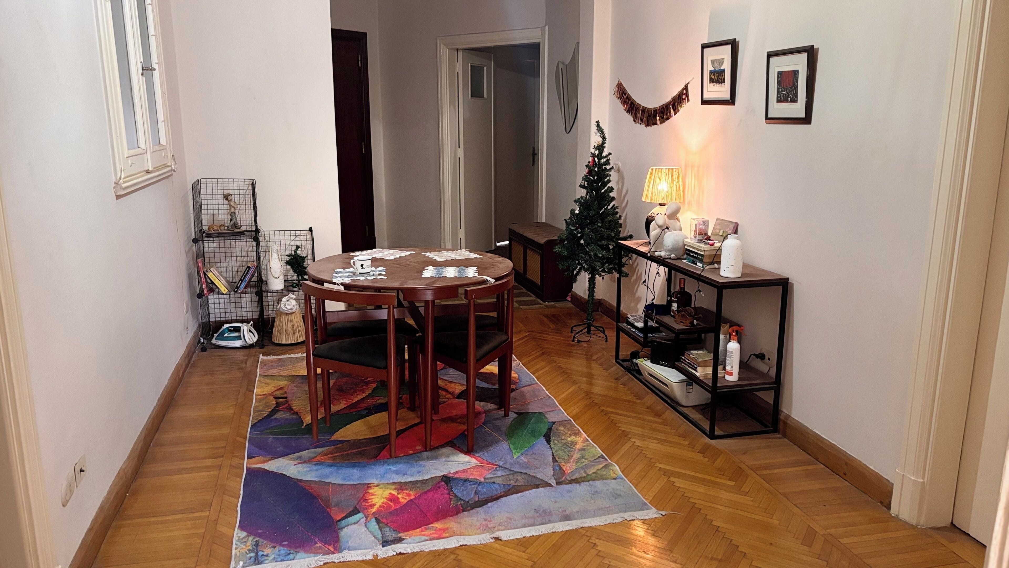 A dining area with a round table, chairs, a Christmas tree, and a side table with a lamp.