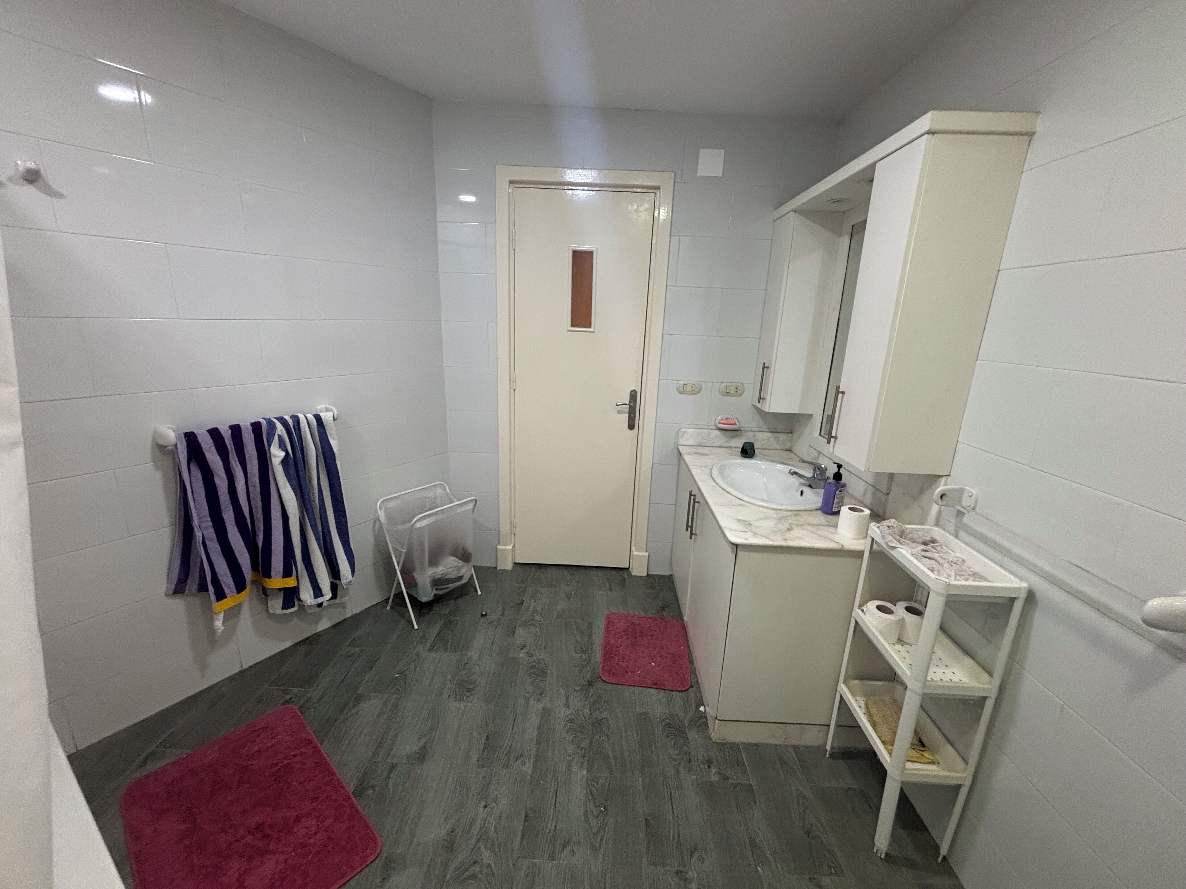 A bathroom with white tiled walls, a wooden floor, a white sink with a mirror above it, a towel rack with striped towels, and a red rug on the floor.