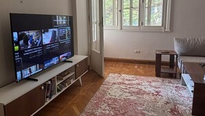 A living room with a flat-screen TV on a stand, a sofa, a small table, and a large window with a view of greenery. - Quiet Spacious Room.. Garden City, 2 Min to Nile (Cairo Governorate)