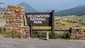 Puinen kyltti, jossa lukee ”Yellowstone-kansallispuisto”, on sijoitettu kivimuurin ympäröimäksi, ja sen edessä on metallinen portti.
