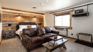 A bedroom with a brown leather sofa, a wooden coffee table, a bedside table with a lamp, and a window with blinds. - Escape to The Adventure Inn Yellowstone- Absaroka, just a minute from the park! (West Yellowstone)