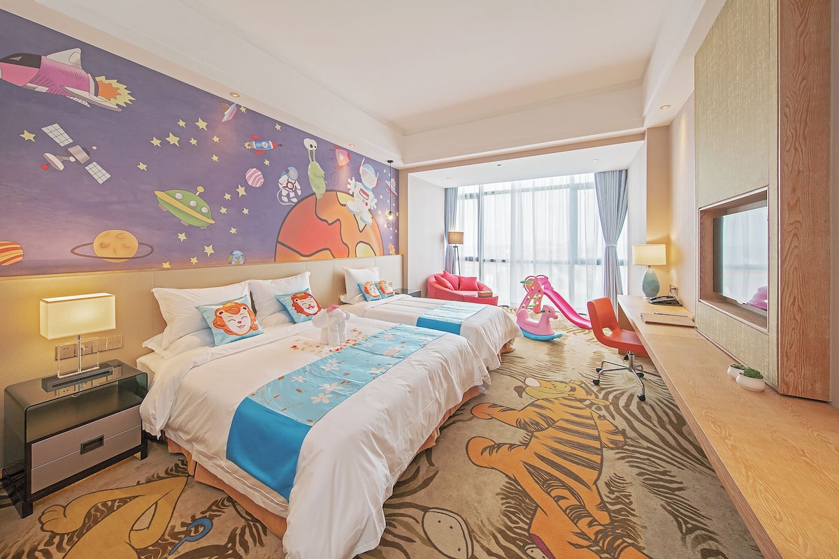 A hotel room with two beds, a large mural of space-themed artwork on the wall, a large window with curtains, and a tiger-patterned carpet.