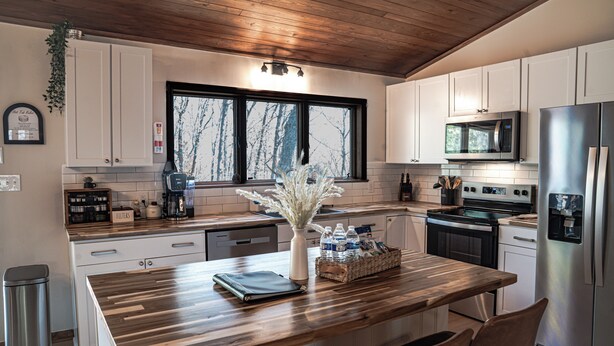 A modern kitchen with white cabinets, a wooden island, stainless steel appliances, and a view of trees through the window.