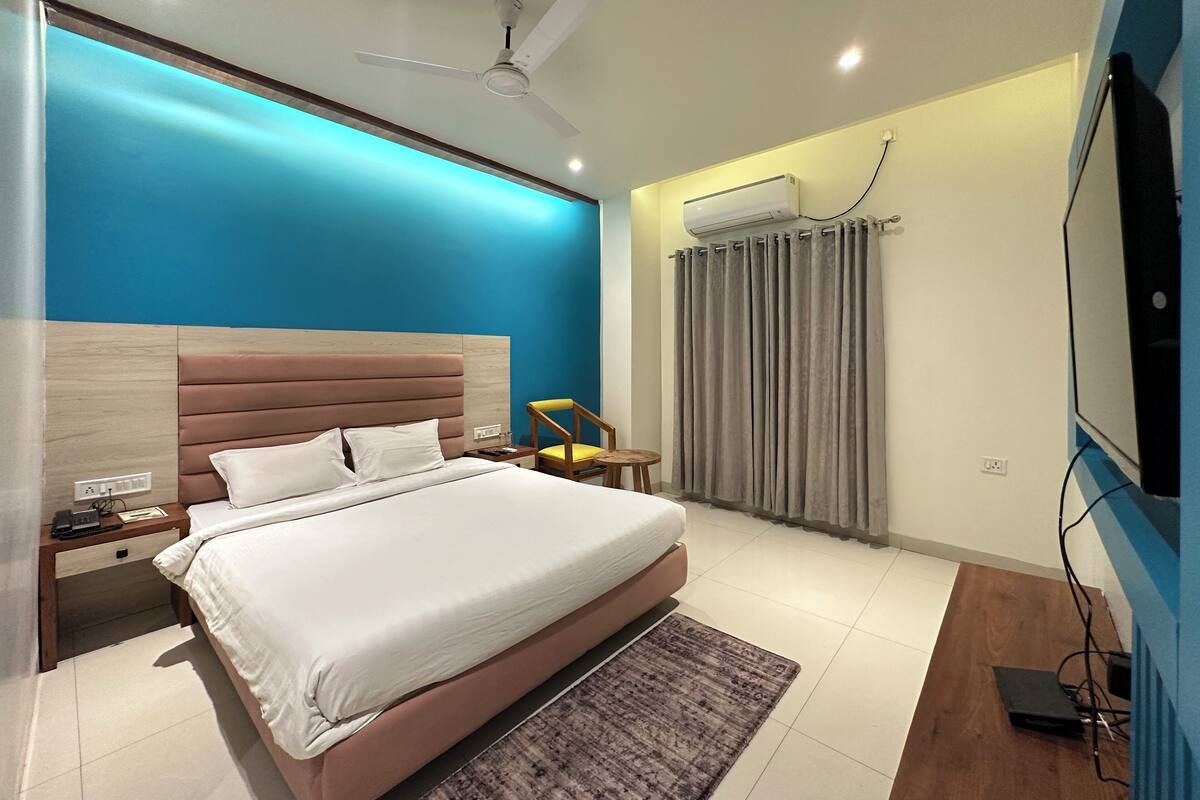 A modern hotel room with a large bed, a bedside table with a phone, a chair, a TV, and an air conditioning unit.