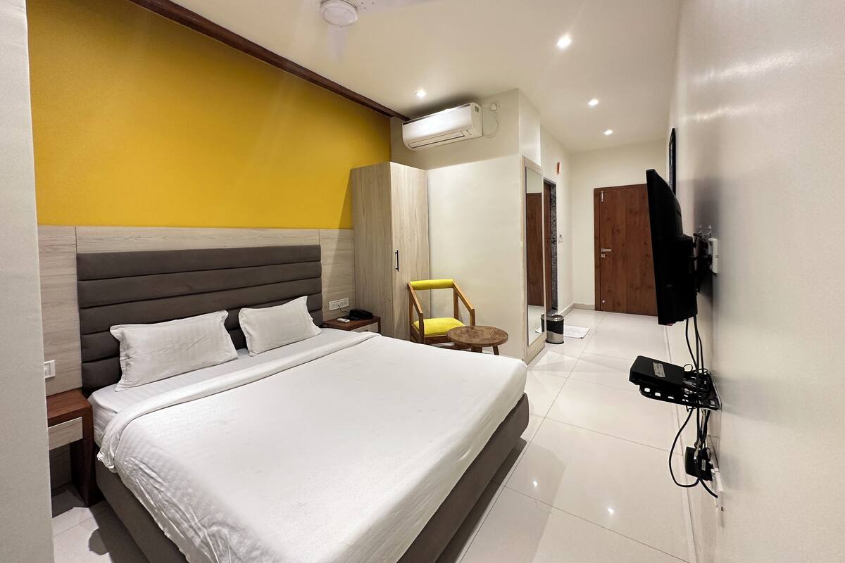 A hotel room with a bed, a bedside table, a chair, a TV mounted on the wall, and an air conditioning unit.