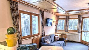 A cozy room with a sofa, a wooden table, and a bench. There are large windows with curtains, a television mounted on the wall, and a radiator.