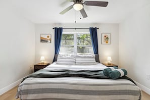Une chambre à coucher comprenant un lit, deux lampes de chevet, un ventilateur de plafond, une fenêtre avec des rideaux et des œuvres d’art accrochées aux murs.