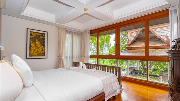 Une chambre dotée d’un grand lit, d’un bureau en bois, d’une fenêtre donnant sur un paysage verdoyant et d’un plafond orné de motifs décoratifs.