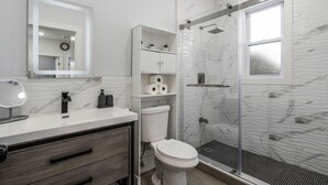 Moderni kylpyhuone, jossa on marmoripintainen seinäosa, lasiseinäinen suihku, wc ja allaskaappi, jossa on peili ja saippua-annostelija.