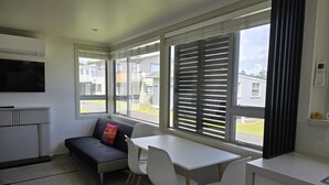 A compact kitchenette with a built-in oven and microwave, a grey sofa with a colorful cushion, a white dining table with two chairs, and large windows with blinds.