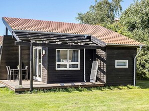 A small, dark wooden house with a tiled roof, a covered patio with chairs, and a window.