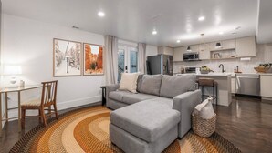 Una sala moderna con un sofá seccional gris, una pequeña mesa de comedor con una silla, y una cocina con armarios y electrodomésticos blancos.