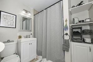 A bathroom with a white sink, mirror, and toilet. There is a shower curtain, a towel rack, and shelves with toiletries.