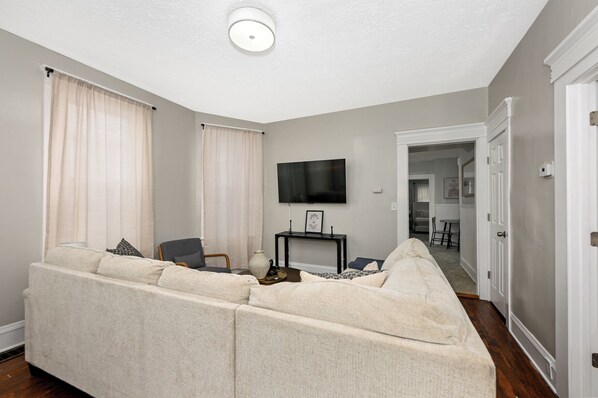 A living room with a beige sectional sofa, a small table with a flat-screen TV, and a chair. There is a door leading to another room.